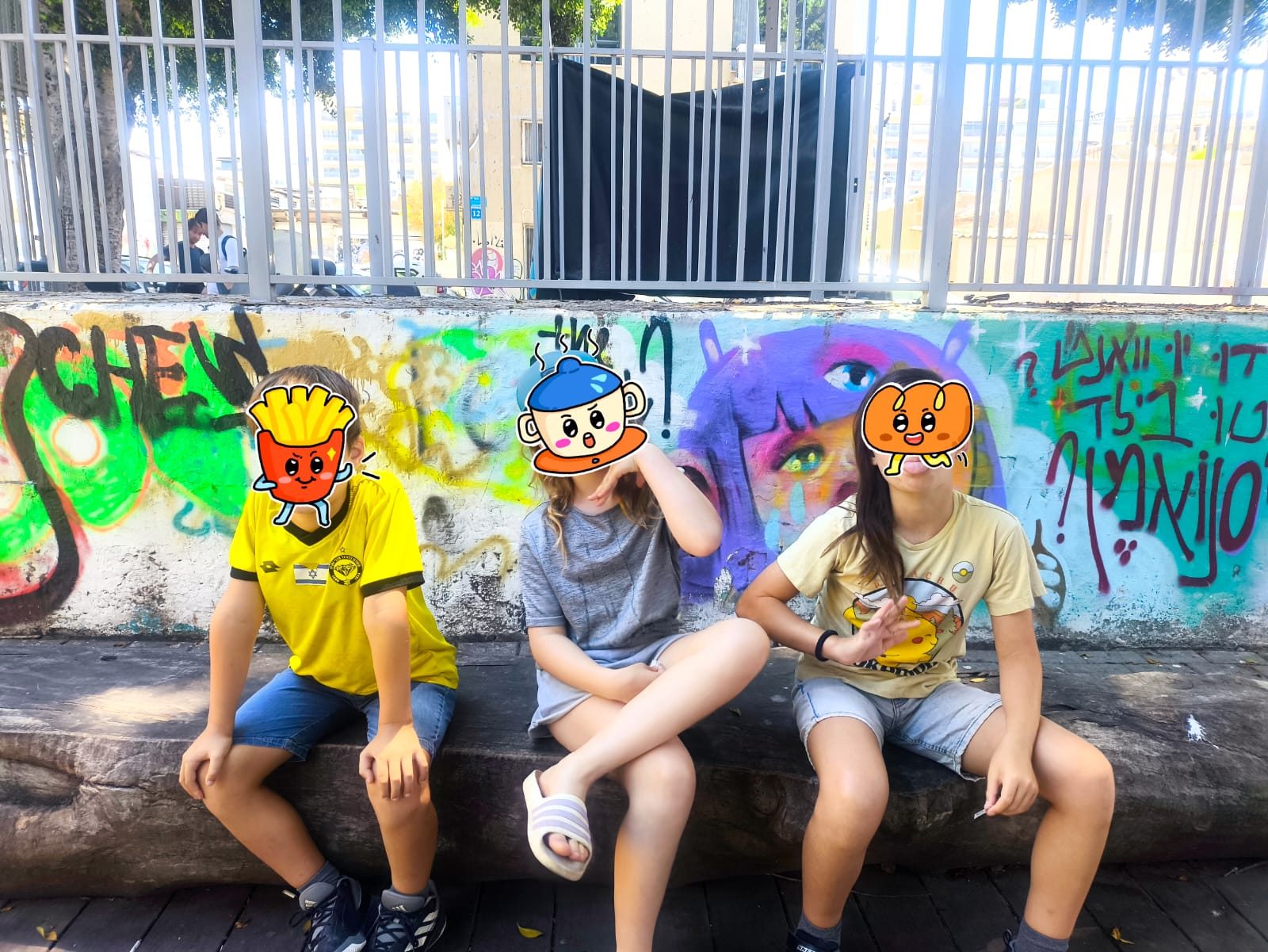 שלושה ילדים יושבים על ספסל מול קיר גרפיטי צבעוני